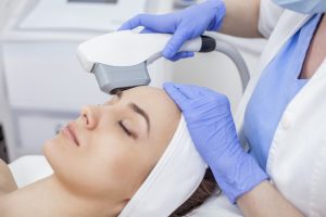 Woman receiving Intense Pulsed Light (IPL) treatment at a skincare clinic, with a technician applying the device on her forehead, highlighting aesthetic and wellness services.
