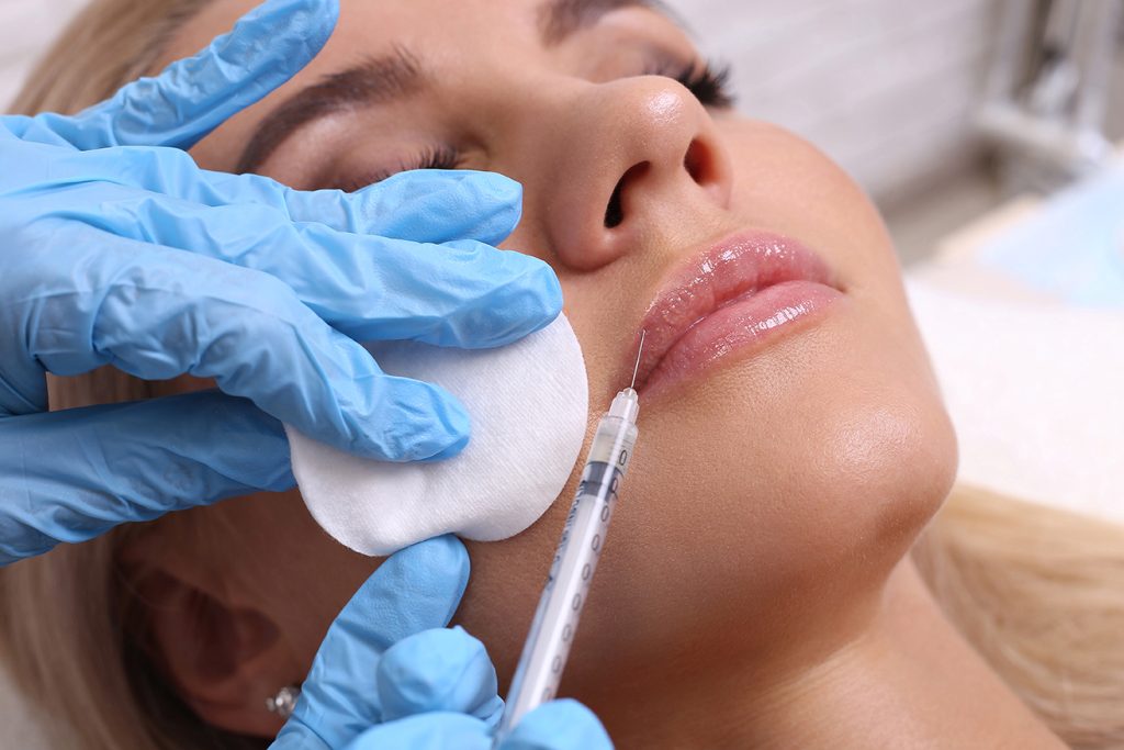 Woman receiving lip injections with a syringe while a clinician holds a cotton pad, showcasing a cosmetic procedure at a medical spa.
