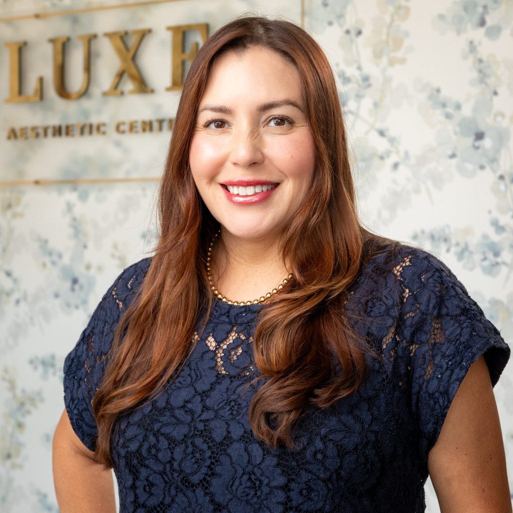 Clarissa Semeniuk, Board Certified Family Nurse Practitioner at Luxe Aesthetic Center, smiling in front of a floral backdrop and Luxe Aesthetic Center sign.
