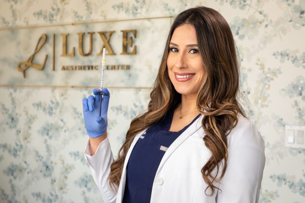 Aesthetic professional holding a syringe, promoting lip fillers and injectable treatments at Luxe Aesthetic Center, with floral wallpaper in the background.